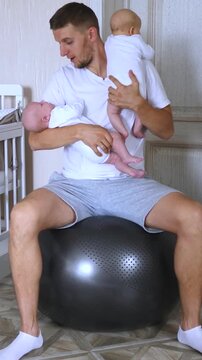 Young dad sitting on a fitness ball and gently rocking his two adorable newborn twins to sleep, demonstrating paternal love and care in a modern family setting in their nursery room