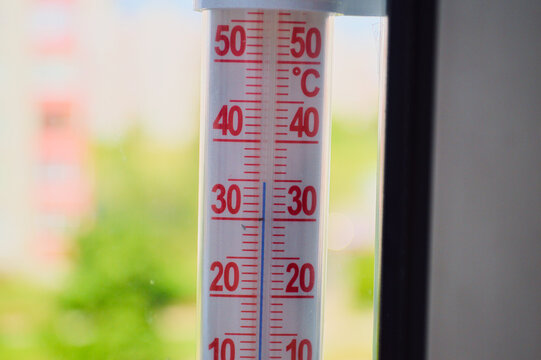 Close-up of a white outdoor thermometer with red Celsius scale markings showing around 28 degrees on a bright sunny summer day with green foliage