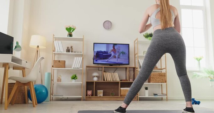 Woman doing home workout with online fitness training. She stretches and practices yoga in a room, following a trainer on the television with a mat and dumbbells. Home fitness and wellness idea