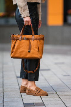 Brown leather handbag carried by stylish woman wearing beige fuzzy coat and black tailored pants while walking on busy city street with blurred urban backdrop and neutral fashion colors