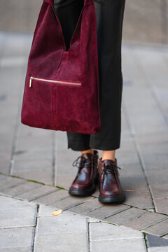Burgundy suede tote with gold zipper pocket carried by stylish shopper beside black pants and matching lace-up boots on urban sidewalk in city setting, with burgundy, tote, bag, suede detail