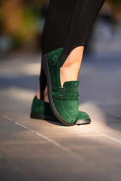 Closeup green suede loafer paired with dark tapered trousers on urban sidewalk, showcasing contemporary street fashion, bold color contrast, visible footwear details, and daylight styling