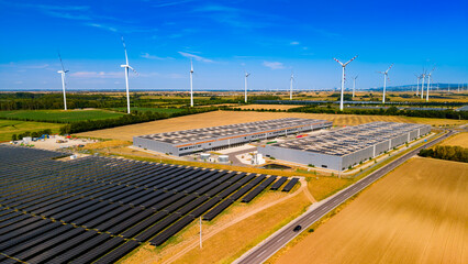 Fototapeta premium Renewable energy sources power a large facility in a rural area. Solar panels and wind turbines are visible beside a large building in an open field. The setting is bright and clear.