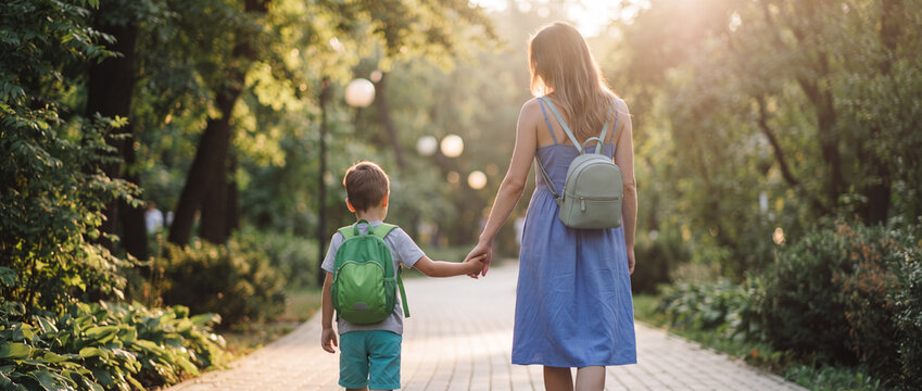 Mother walking with son in sunny park with backpacks holding hands on stone pathway surrounded by lush green trees, shot from behind in warm backlight for family lifestyle concepts