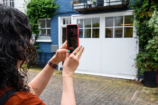 Over-the-shoulder view of a woman with curly hair raising her smartphone to photograph a charming blue and white mews house facade with climbing greenery.