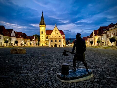 Silhouette of executioner statue on Town Hall Square in Bardejov, Slovakia at night. Illuminated Old Town Hall, St. Giles Basilica, and blue hour sky in UNESCO World Heritage site.