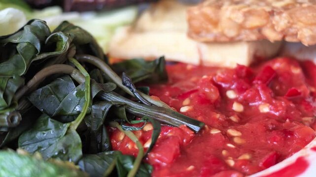 Extreme macro view highlighting the rich texture of crushed red chili sambal seeds alongside boiled cassava leaves, lime, and golden fried soybean tempeh on plate.
