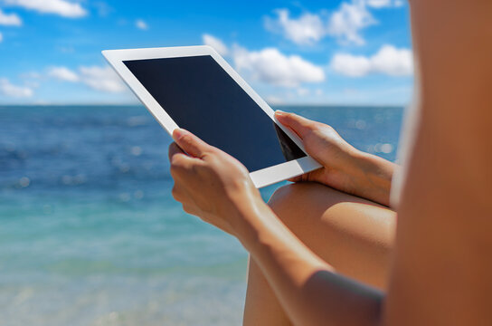 A person holds a digital tablet with a blank screen on a tropical beach.