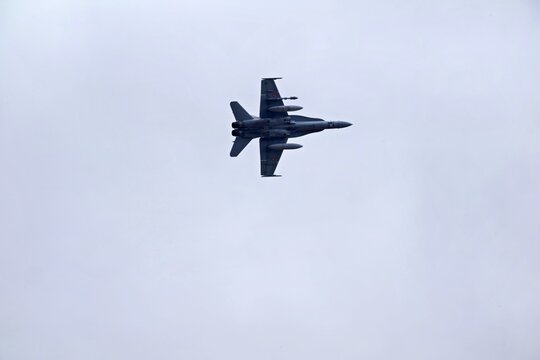 A military fighter jet is flying high in a cloudy sky. The aircraft is captured in an aerial view with gray clouds. It represents modern air defense capabilities.