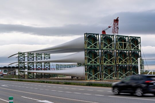Large white wind turbine blades rest on green metal scaffolding towers beside a highway under an overcast sky, with a vehicle passing in the foreground and a red crane visible above the structure