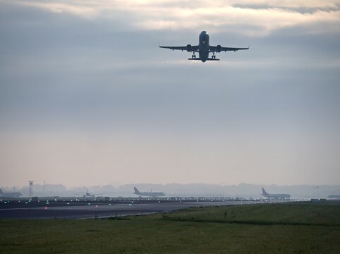 An airplane takes off from the runway at the airport under a cloudy sky. Other aircraft are visible on the runway, with runway lights illuminating the area.