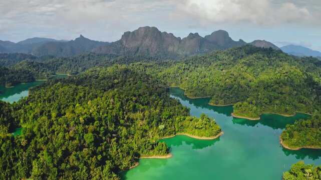 Khao Sok National Park, emerald green water of Cheow Lan Lake surrounded by lush tropical rainforest. Towering limestone mountains and dramatic cloudy sky. Serene nature aerial view landscape panorama