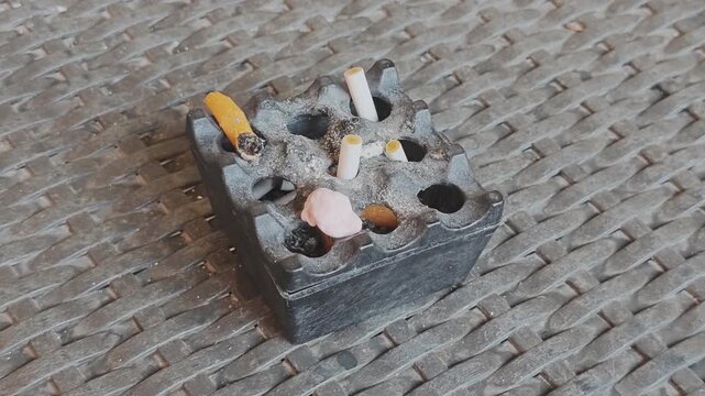 Dirty ashtray full of cigarette butts and ash sitting on a woven patio table. The concept of smoking addiction and bad habits