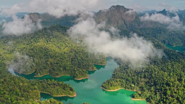 Aerial landscape of KHAO SOK National Park in Thailand, vibrant turquoise lake waters winding through lush green rainforests, limestone mountains shrouded in misty clouds. Drone shote rise up panorama