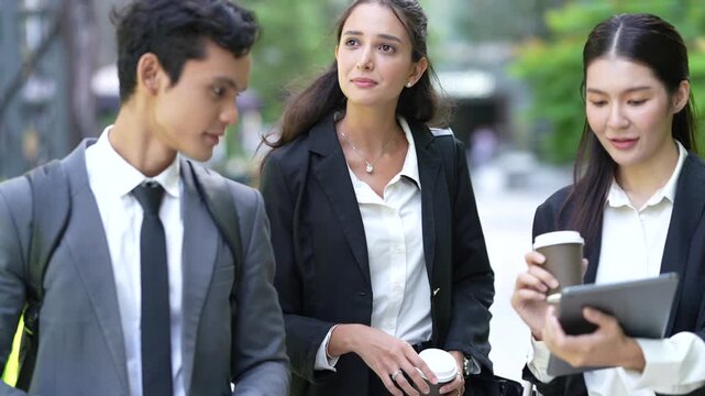 Business colleagues walk side by side sharing a tablet screen and drinking takeaway coffee, gesturing at the display as they move through a landscaped plaza in the city business district