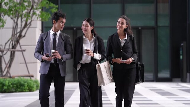 Three business colleagues walk towards camera across a graphic patterned plaza in front of a modern glass office building, holding coffee and phones and talking as the shot pushes in