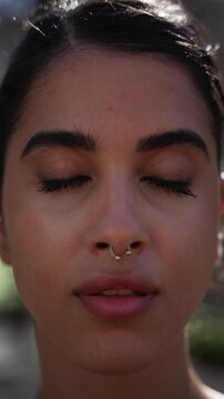 Vertical close-up portrait of a young Iranian woman outdoors with eyes closed in meditation before opening them and smiling at camera, conveying mindfulness, calm energy and emotional wellbeing.