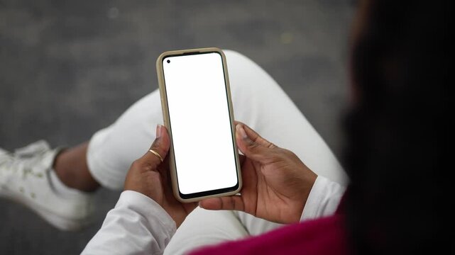 Close up of a relaxed sitter holding a phone with an empty display as the scene conveys casual mobile use. The wearer wears white jeans and a red sweater, seated under gentle light with a calm