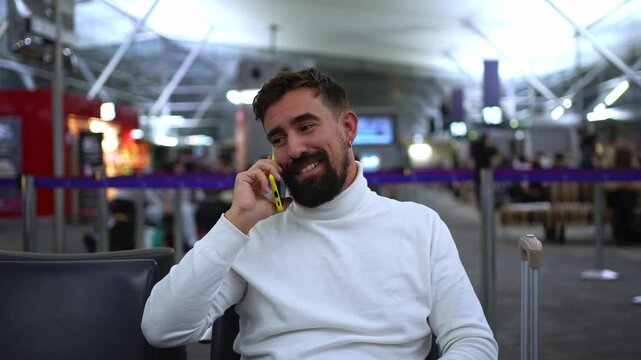 Bearded man smiles while speaking on his smartphone as he sits in an airport terminal waiting for his journey, then checks the phone after the call.