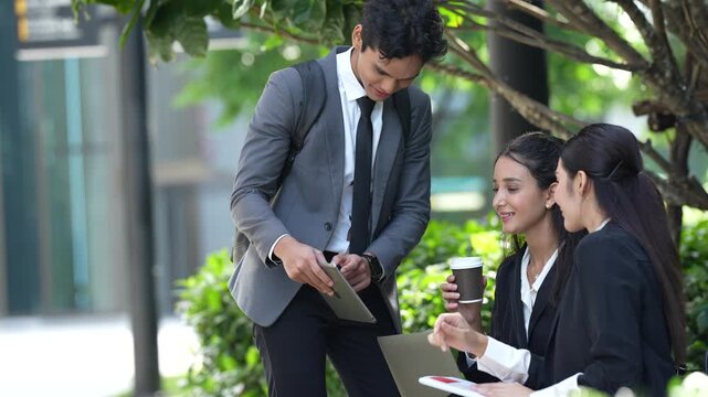 Businessman standing and pointing at a laptop screen while two seated businesswomen listen and sip takeaway coffee during an informal outdoor team meeting in a landscaped office plaza