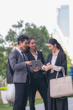 A diverse team of three business professionals comparing notes over a tablet screen in a green terrace with towers in the background, a modern picture of teamwork in a green city district