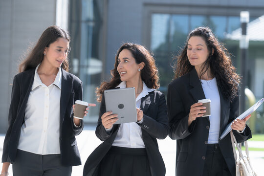 Three female colleagues in matching black business suits, talking as they walk in step, cross a plaza on the way to a client meeting with a white tote bag and printed papers.