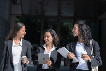 Three female colleagues in matching black business suits, smiling in the bright daylight, move as a group across a sunlit forecourt with a white tote bag and printed papers. © ultramansk
