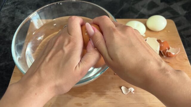 Woman's hands peeling the shell from a boiled egg over a bowl, home cooking, food preparation, kitchen routine, healthy protein snack.