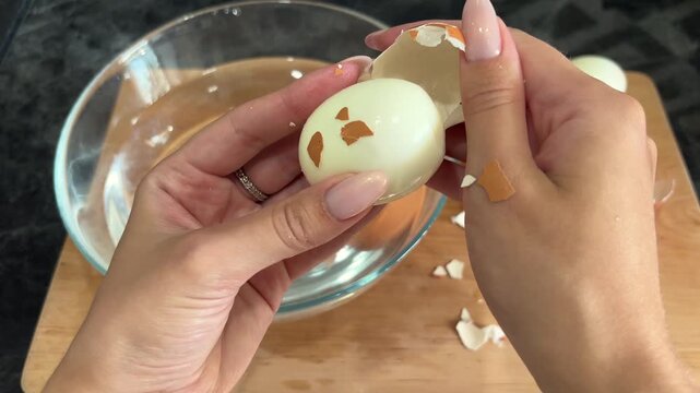 Close view of a woman peeling a boiled egg by hand over a bowl, healthy protein food, simple cooking, kitchen preparation, everyday meal routine.