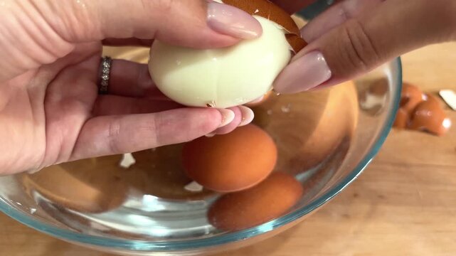 Woman holding a peeled boiled egg above a bowl of eggs, home cooking, food preparation, protein snack, kitchen routine, simple meal.