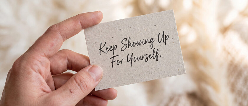 Close up of a hand holding a small card with the inspirational quote keep showing up for yourself written on it