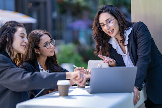 A diverse team of three Hispanic businesswomen in dark suits, deep in an animated discussion, sketch out an idea with a stylus on a tablet pointing at the laptop screen.