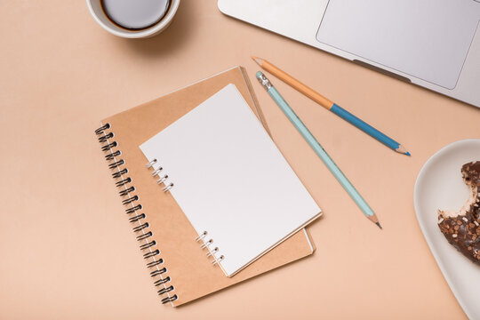 Notebook and writing tools on a desk with coffee and dessert beside a laptop on a light surface