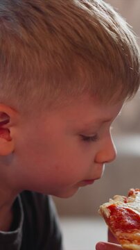 Close up of young boy eating pepperoni pizza holding slice with focused expression. Pizza details visible including melted cheese and red sauce on crust. Soft indoor lighting with blurred