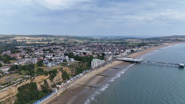 Sandown, United Kingdom - 21 June 2025: Aerial view of Sandown Pier extending into the English Channel, with the coastal town of Sandown, sandy beaches, and cliffs under a cloudy sky.
