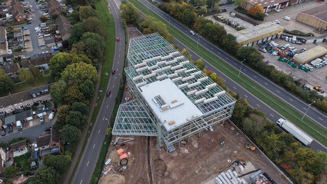 Aerial view of a warehouse construction site featuring a steel framework and partial roof covering, situated near a road and residential area in Cheshunt, England, United Kingdom.