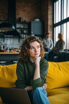 Woman with dark, curly hair in a green sweater and jeans, seated on a vibrant yellow sofa with a laptop, in a large co-working space.