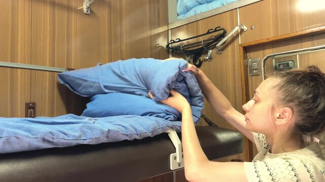 Young passenger folding and rolling bedding in a sleeper train compartment, rail travel routine, overnight journey, practical travel lifestyle.