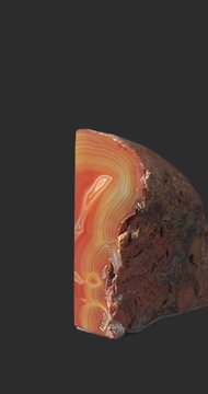 Polished agate specimen rotating against a dark gray background, revealing translucent red, orange and brown bands, concentric mineral patterns and a glossy polished surface.