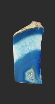 Vertical view of a polished blue and white banded agate specimen rotating on a dark gray background, revealing concentric patterns, smooth cut surfaces and a rough natural exterior.