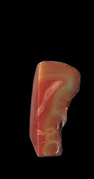 Polished agate specimen rotating against a dark background, revealing red, orange and brown concentric bands and translucent mineral layers across its smooth cut surface.
