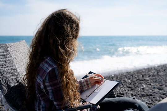 Unrecognizable woman making notes in a notebook by the sea in sunny weather. Casual coastal lifestyle scene representing reflection, mindfulness, creativity and personal time.