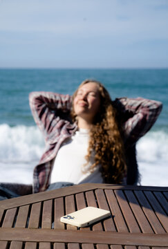 Young woman relaxing by the sea in warm sunlight, resting in a beach chair and taking a break from her phone and everyday digital distractions. Concept of digital detox, self-care and slow living.