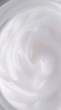 White Cream Swirling. Thick glossy cream moves in smooth folds, creating a soft abstract cosmetic texture.	