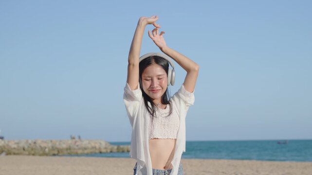 Happy young woman enjoying music and dancing on beach wears wireless headphones, joyful Asian female tourist relaxes near tropical sea during summer vacation under blue sky.