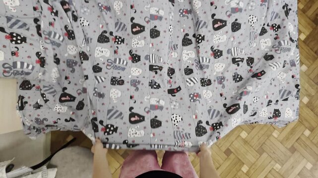 POV hands unfolding grey fabric with cute cartoon cats print on parquet floor.