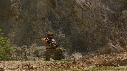 Naklejka premium A special forces soldier in combat gear kneels with a rifle at the base of a cliff, maintaining vigilance during a tactical operation in a rugged environment.