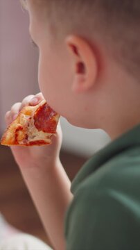 Close up of young boy eating pepperoni pizza. Boy holds slice with visible cheese and tomato sauce bringing it to mouth in casual indoor setting. Soft lighting highlights details of food