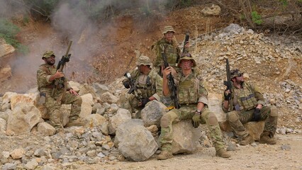 Fototapeta premium An elite team of special forces soldiers in full tactical gear pose together with their rifles amidst smoke and rocks during a military operation in a rugged combat zone.