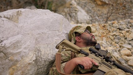 Naklejka premium An exhausted soldier with face paint rests behind a rock, holding his rifle and catching his breath during a tactical military mission in a rugged environment.